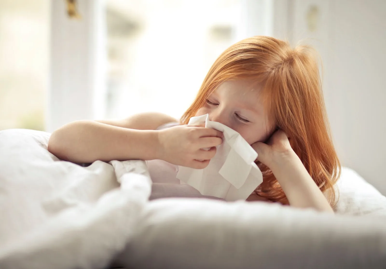 Small sick redhead kid with eyes closed cleaning nose with white tissue paper while lying alone in bed in light modern bedroom in daytime