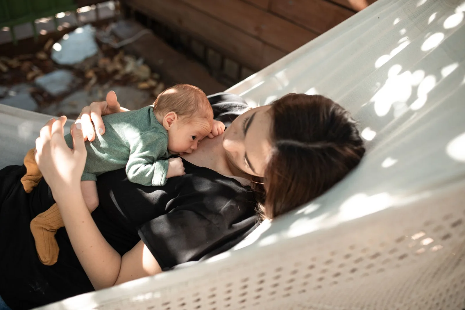 A mother gently holds her newborn baby while lying together in a hammock under sunlight.