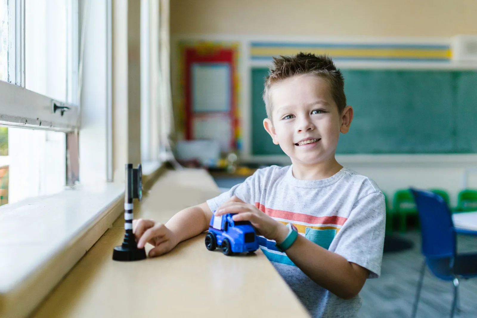 Young boy playing with a blue toy truck at a classroom table by a window.