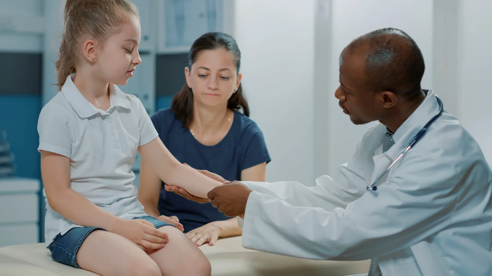 Pediatrician examines young girl's arm while mother observes in clinical setting.