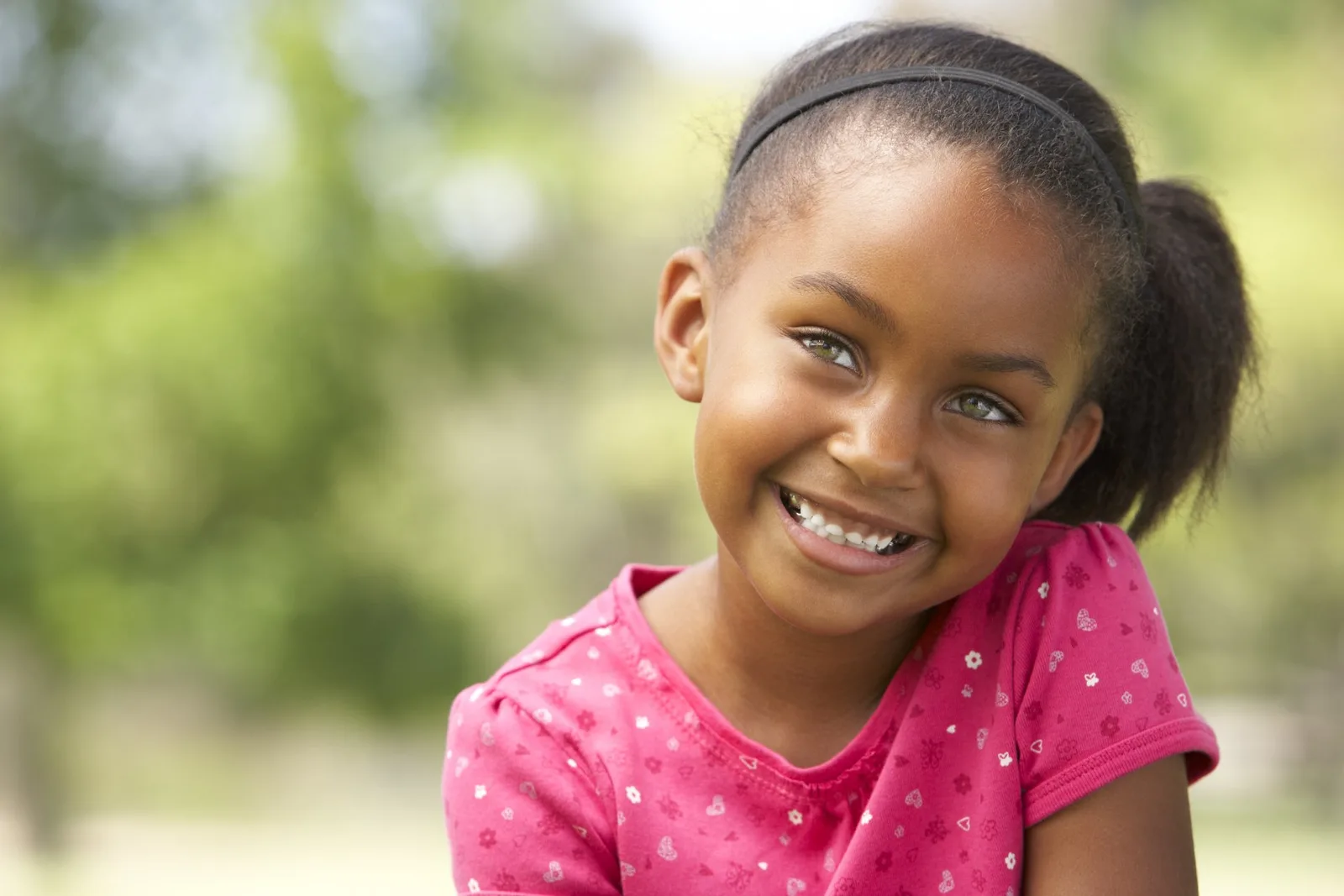 Young girl smiling at camera outdoors wearing pink patterned shirt with blurred green background.