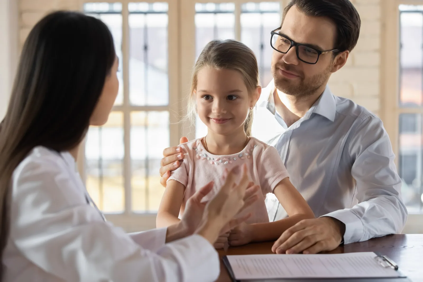 Family meeting with pediatrician in bright office with windows; child smiling during consultation.