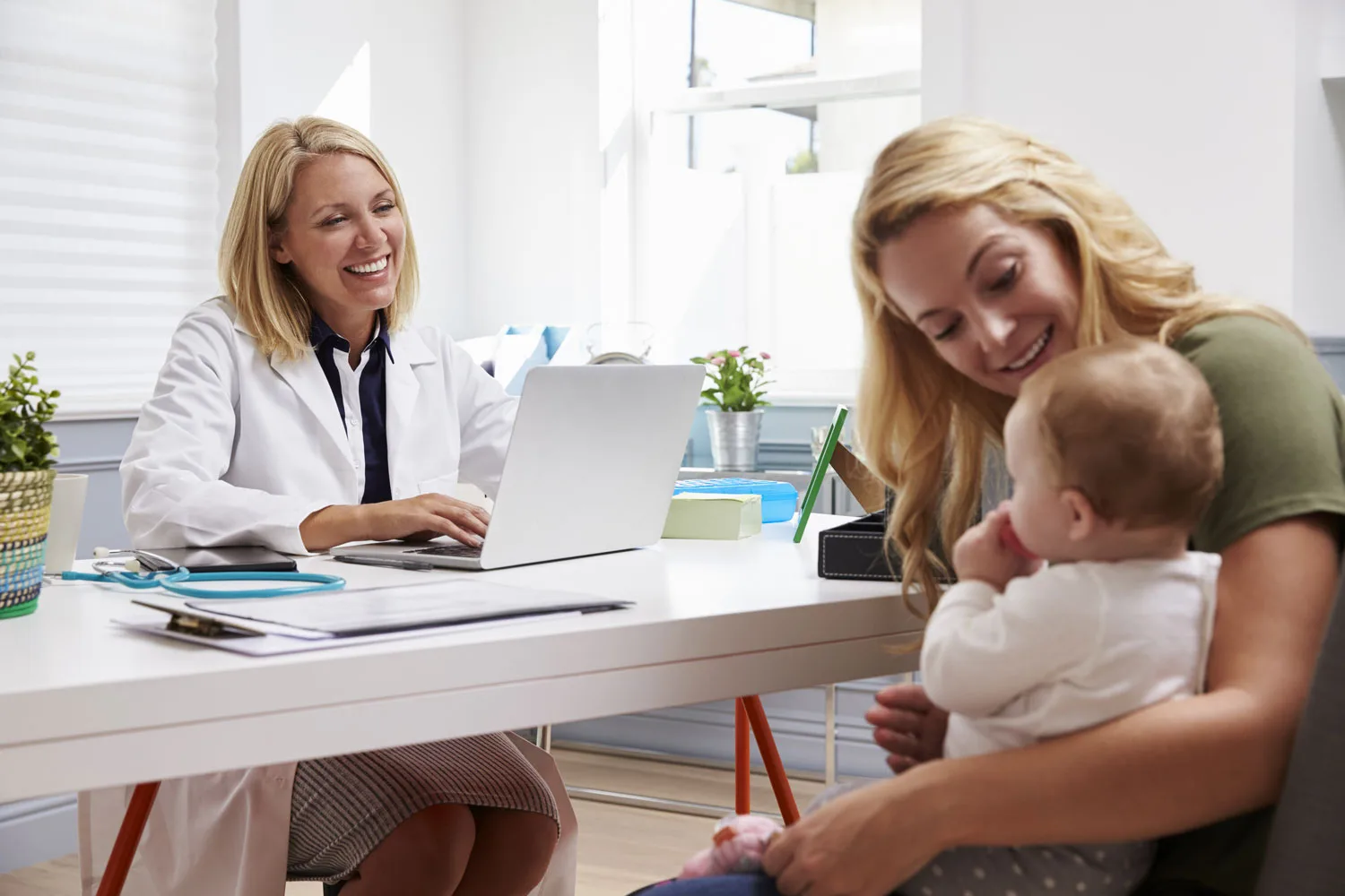 Female pediatrician in white coat smiling at mother holding infant during office visit.