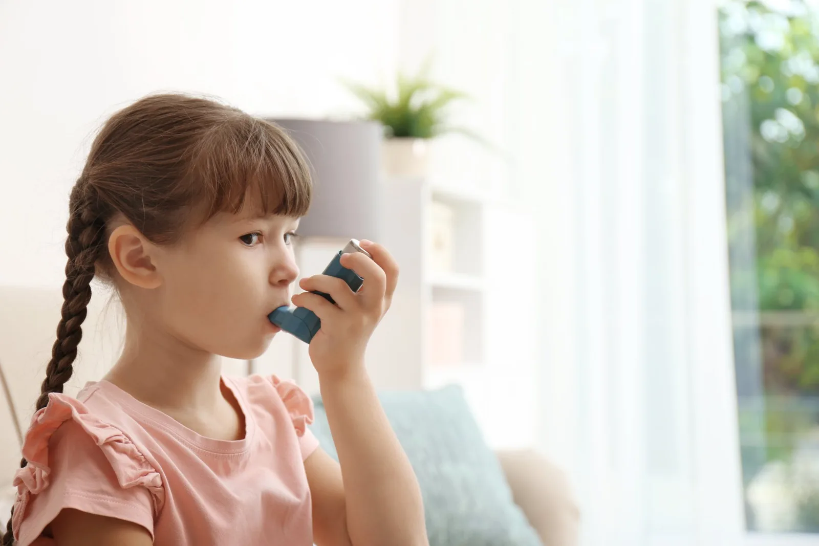 Young girl using blue asthma inhaler indoors, side profile view.