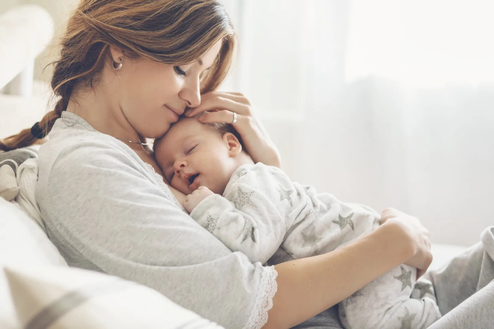 Mother holding sleeping infant in soft lighting, both wearing light colored clothing.