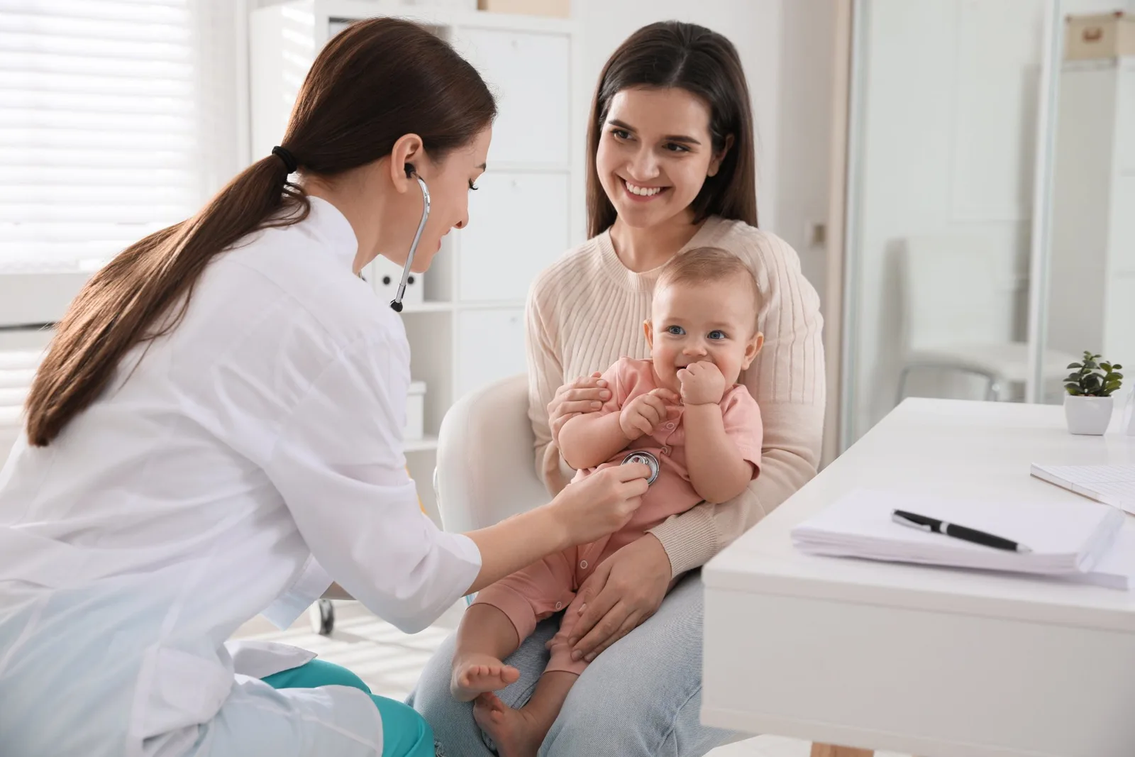 Pediatrician examining infant with stethoscope while mother holds baby in clinic examination room.