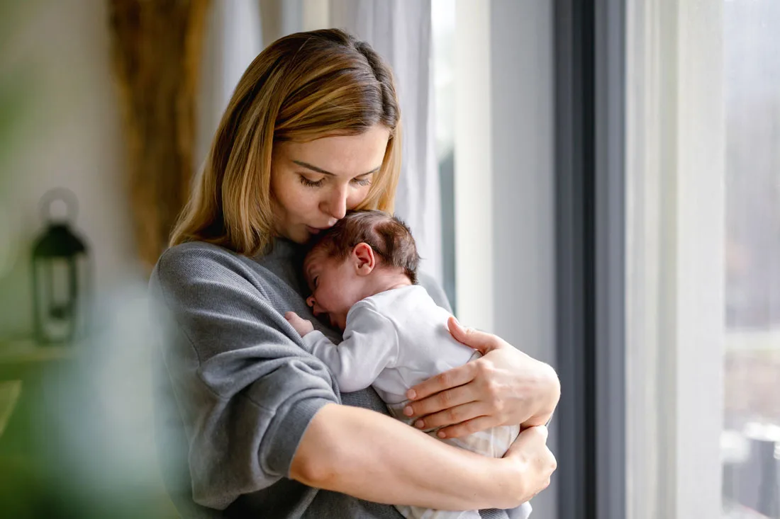 Mother holding newborn infant in gray sweater by window indoors.