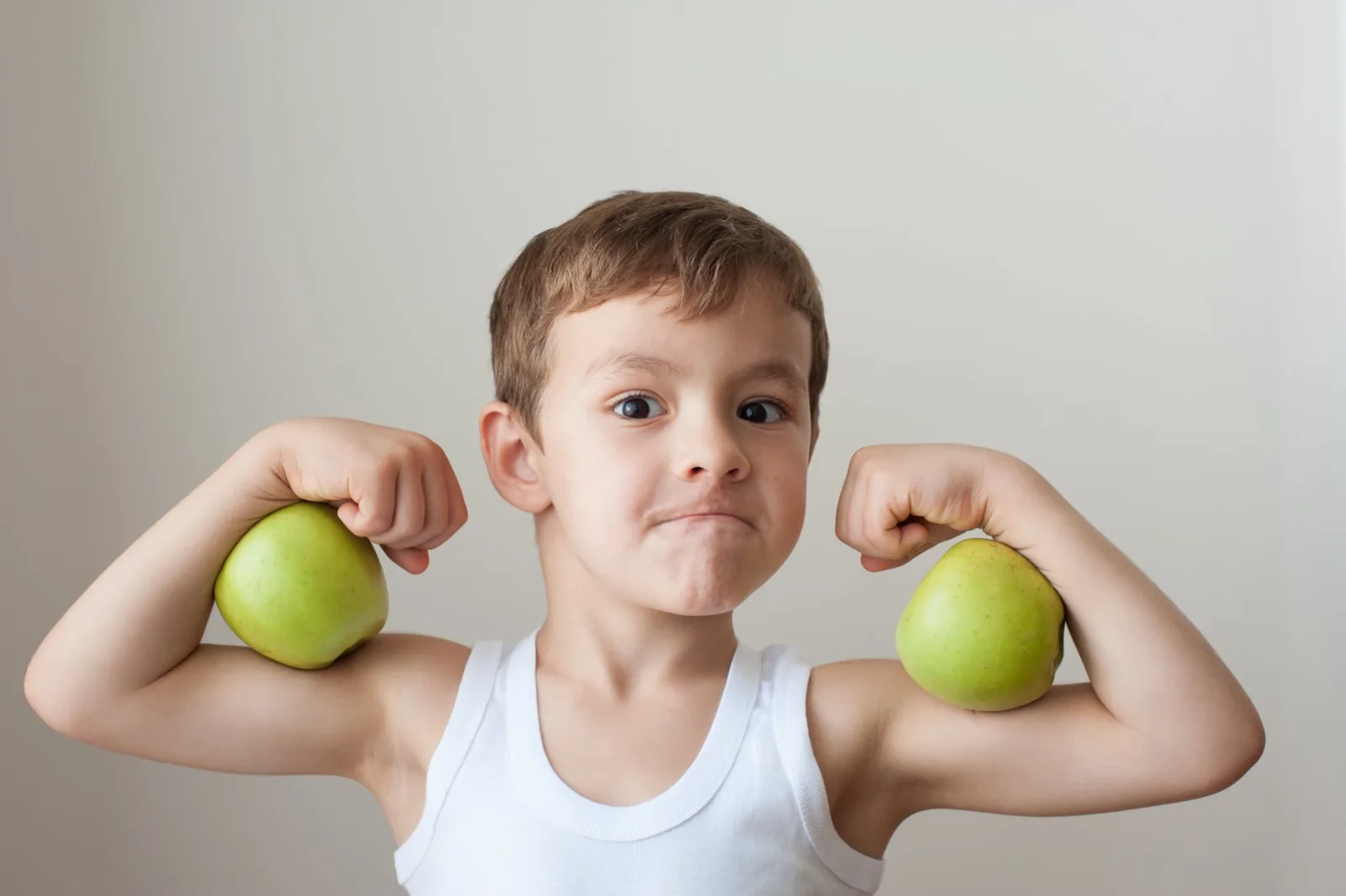 Young boy flexing arms while holding green apples, wearing white tank top.