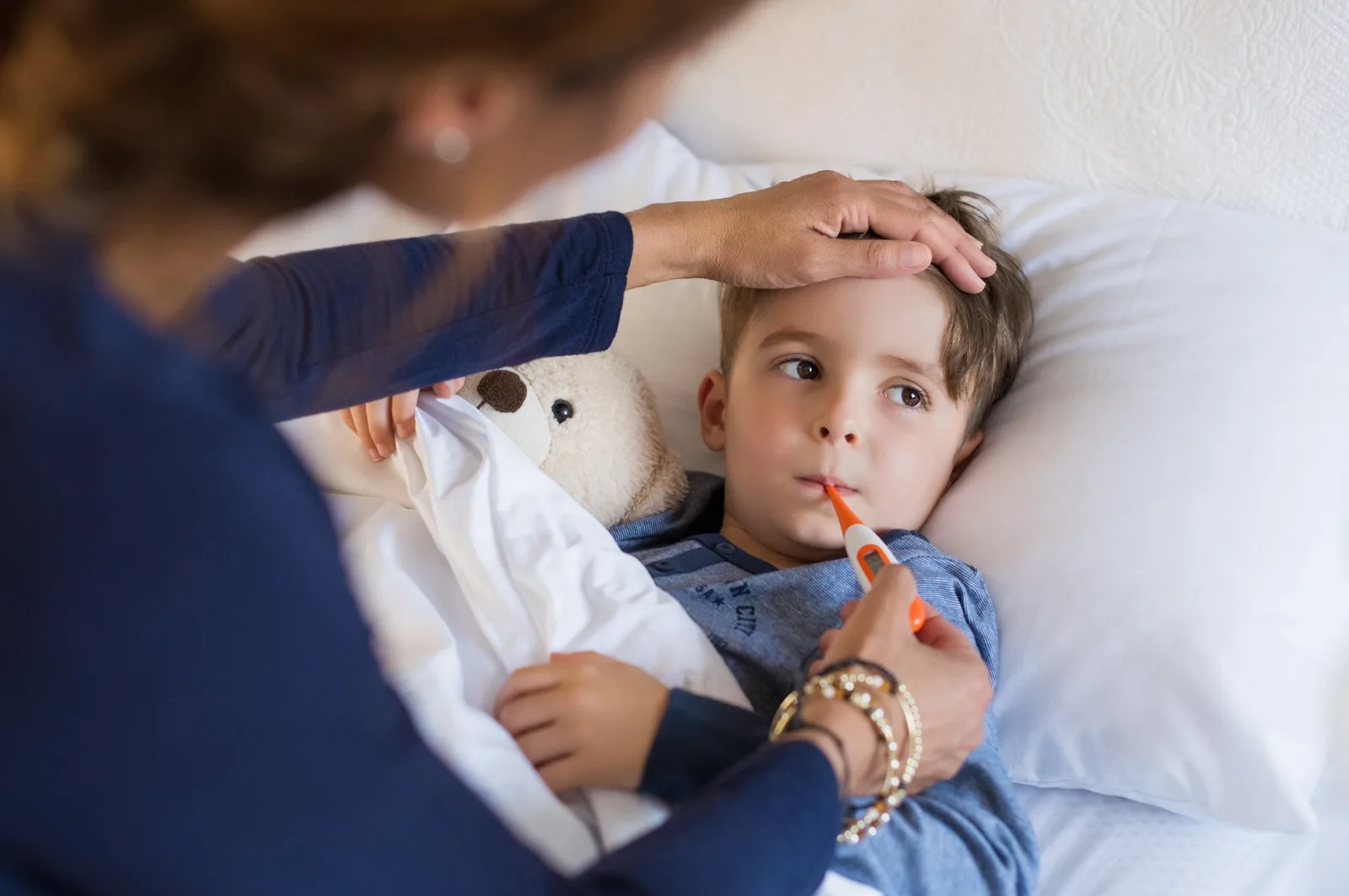 Parent checking sick child's forehead temperature with thermometer in mouth.