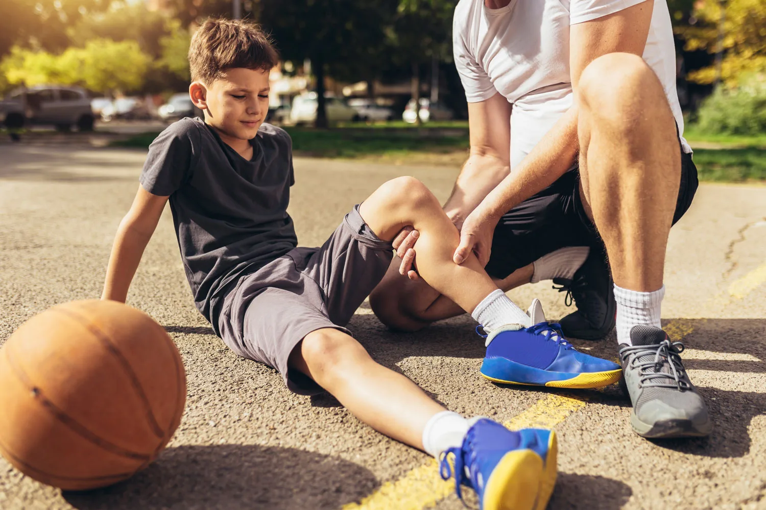 Adult examining child's knee injury on basketball court outdoors.