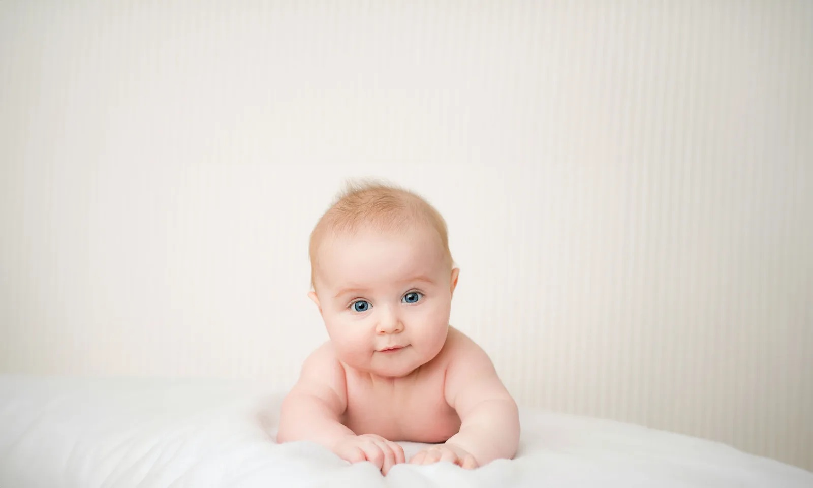 Smiling baby with light hair and blue eyes on white bedding against neutral background.