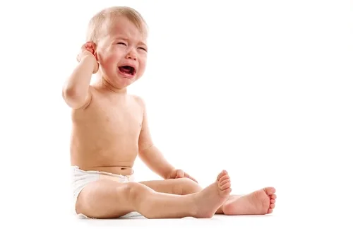 Shirtless toddler sitting, smiling with hand near ear against white background.